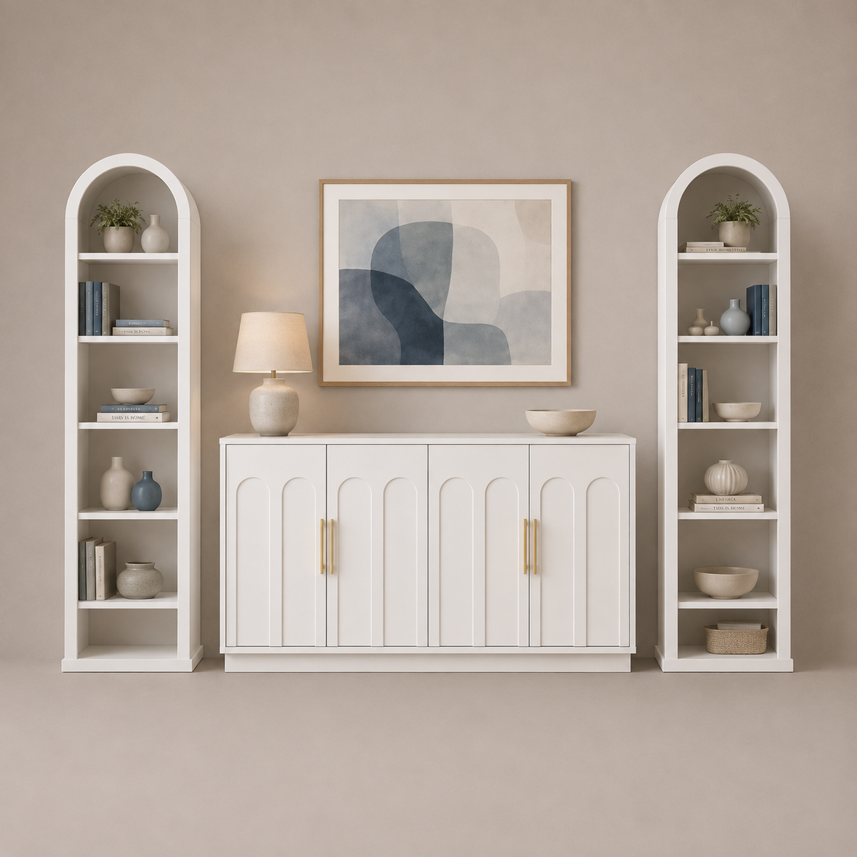 A styled interior scene showing a white sideboard cabinet centered beneath a framed abstract print, flanked by two matching white arched shelving units. The furniture is photographed in a warm neutral room with beige walls and floor, with decorative books, vases, bowls, and a table lamp on the shelves and cabinet.