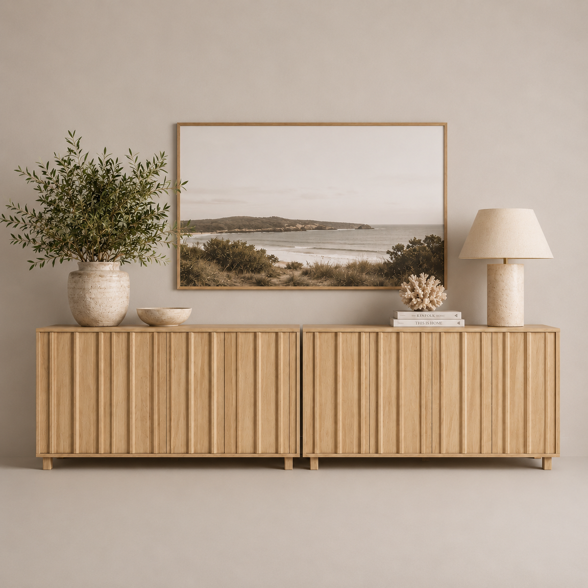 A light natural wood sideboard with vertical slat detailing, styled in a minimalist room setting with a framed coastal landscape print, a potted olive branch, a bowl, a lamp, and books.